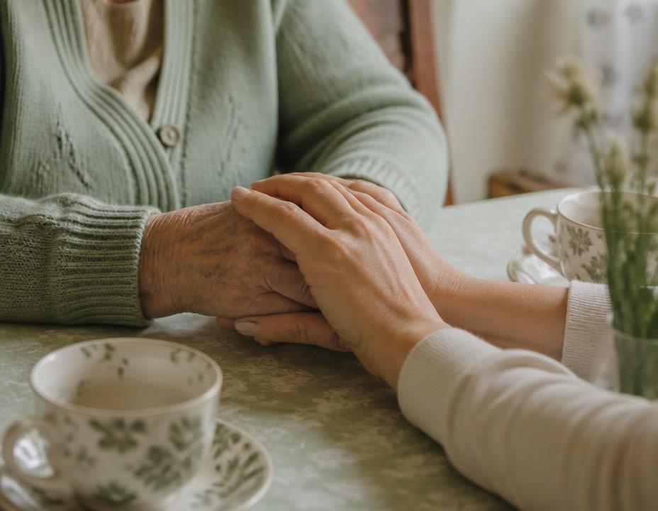 A younger woman holding an older woman's hands across a kitchen table over tea